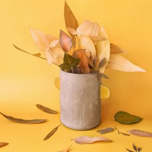 Natureza-morta de outono com folhas secas dispostas em um vaso de cerâmica sobre uma superfície amarela quente. Imagem sazonal aconchegante para papéis de parede, branding de outono, pôsteres, design web, visuais para redes sociais e uso editorial.